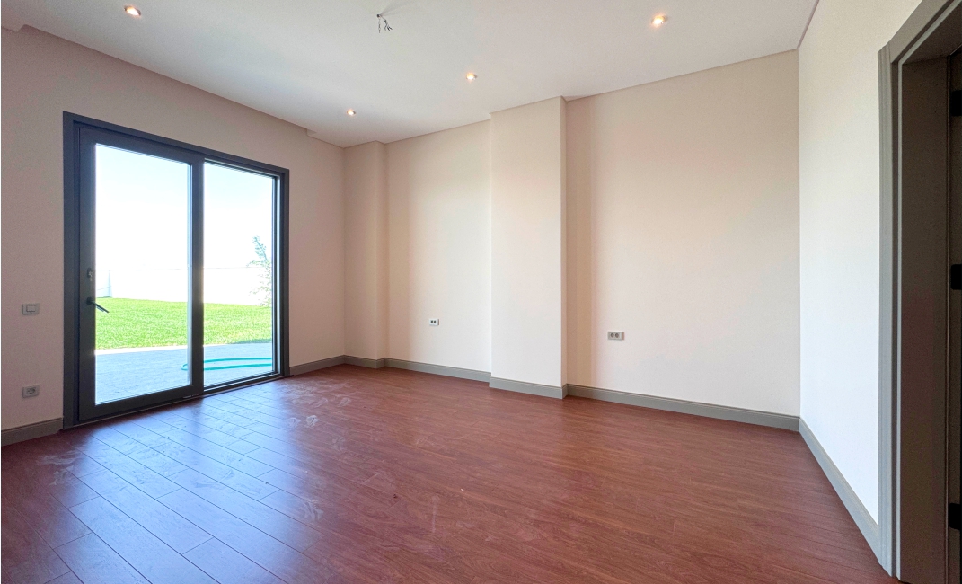 اتاق خواب خالی با کفپوش لمینت چوبی تیره، دیوارهای کرم‌رنگ و درب کشویی شیشه‌ای با دسترسی به حیاط.