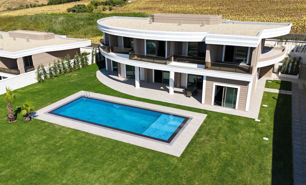 نمای هوایی از یک ویلای لوکس با معماری منحنی، دارای استخر اختصاصی بزرگ و چمن‌کاری وسیع در حیاط پشتی.