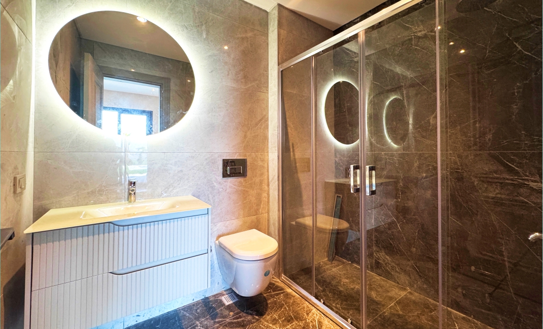 دکوراسیون سرویس بهداشتی مدرن با کاشی‌های طرح سنگ تیره، کابین دوش شیشه‌ای و آینه گرد بک‌لایت (نورپردازی شده).
