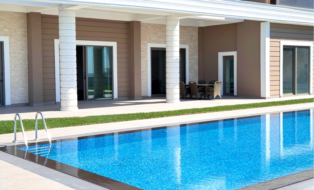 نمای نزدیک از آب استخر و محوطه پاسیو با ستون‌های سفید و میز و صندلی ناهارخوری در فضای باز.