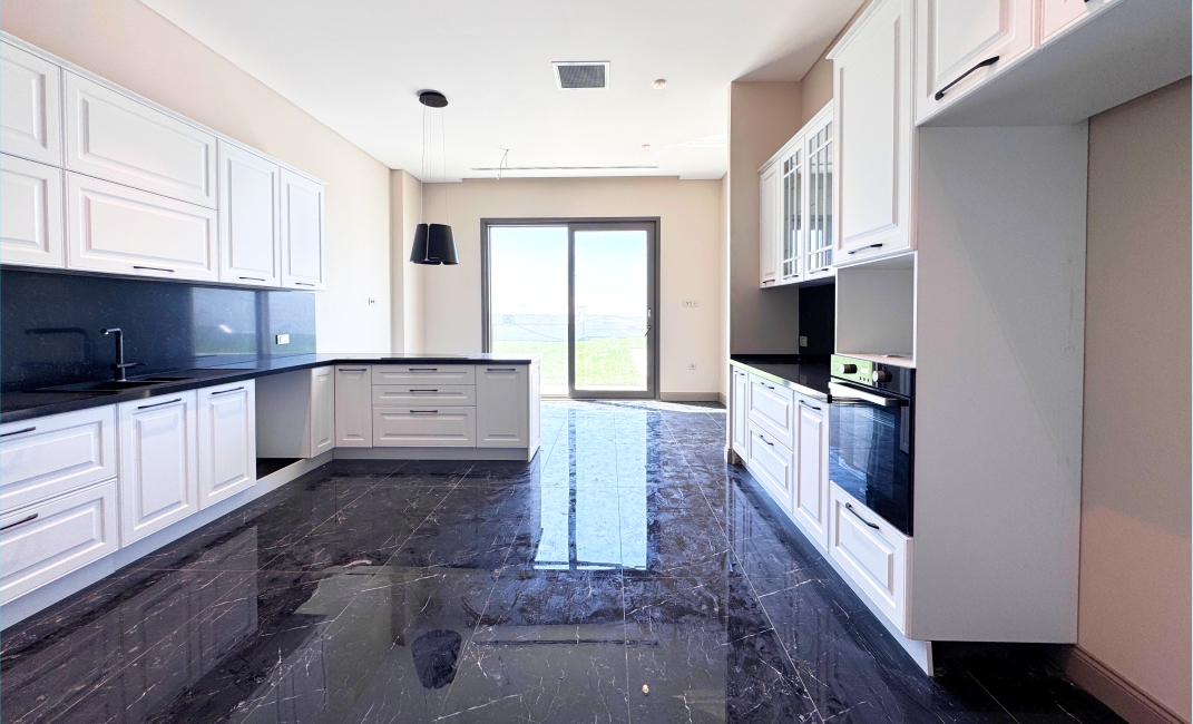 آشپزخانه بزرگ و نورگیر با کابینت‌های سفید کلاسیک، کفپوش سنگ سیاه براق و فضای باز.
