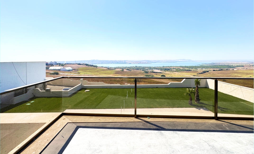 چشم‌انداز پانوراما از تراس پشت‌بام ویلا با نرده‌های شیشه‌ای، رو به دشت سرسبز و دریاچه در دوردست.